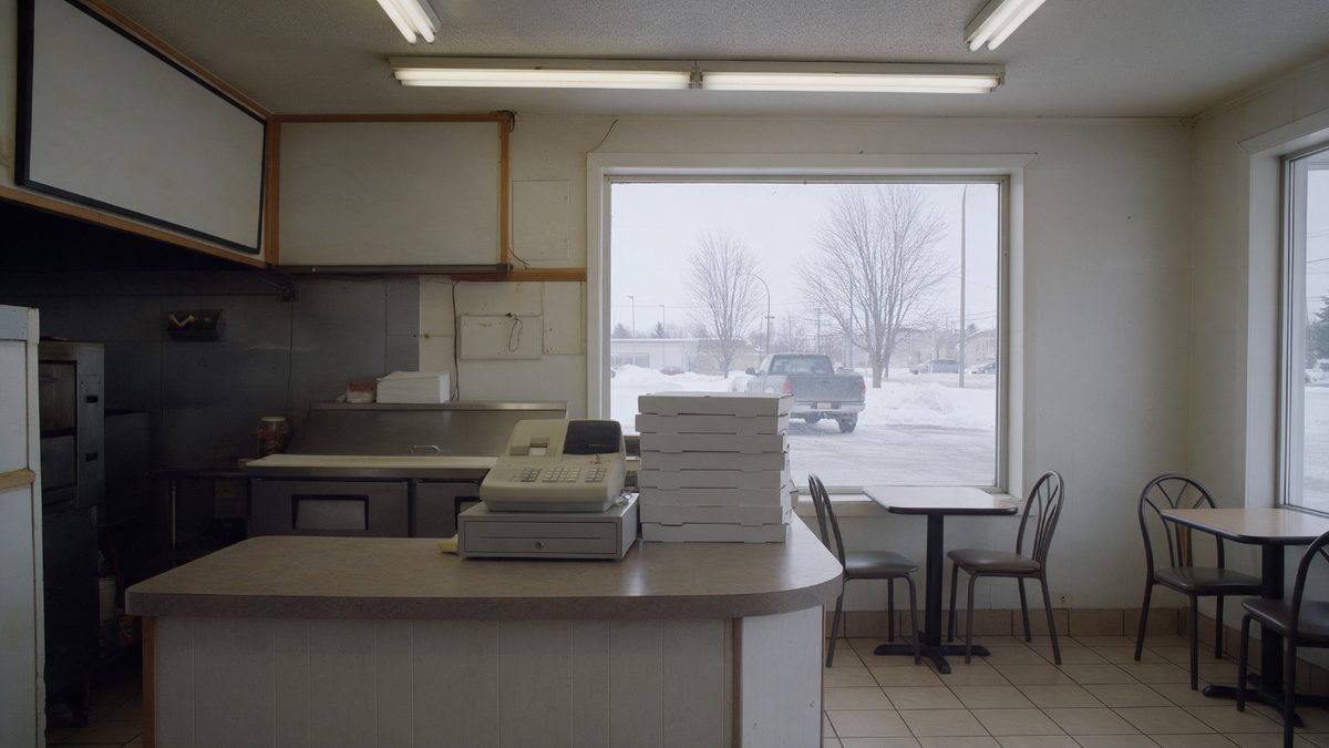 The inside of a small-town takeout counter, with a service counter, stacked pizza boxes and two tables at the window
