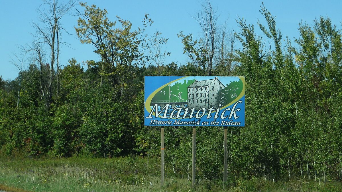 The 'Historic Manotick on the Rideau' sign at the village entrance on a summer day